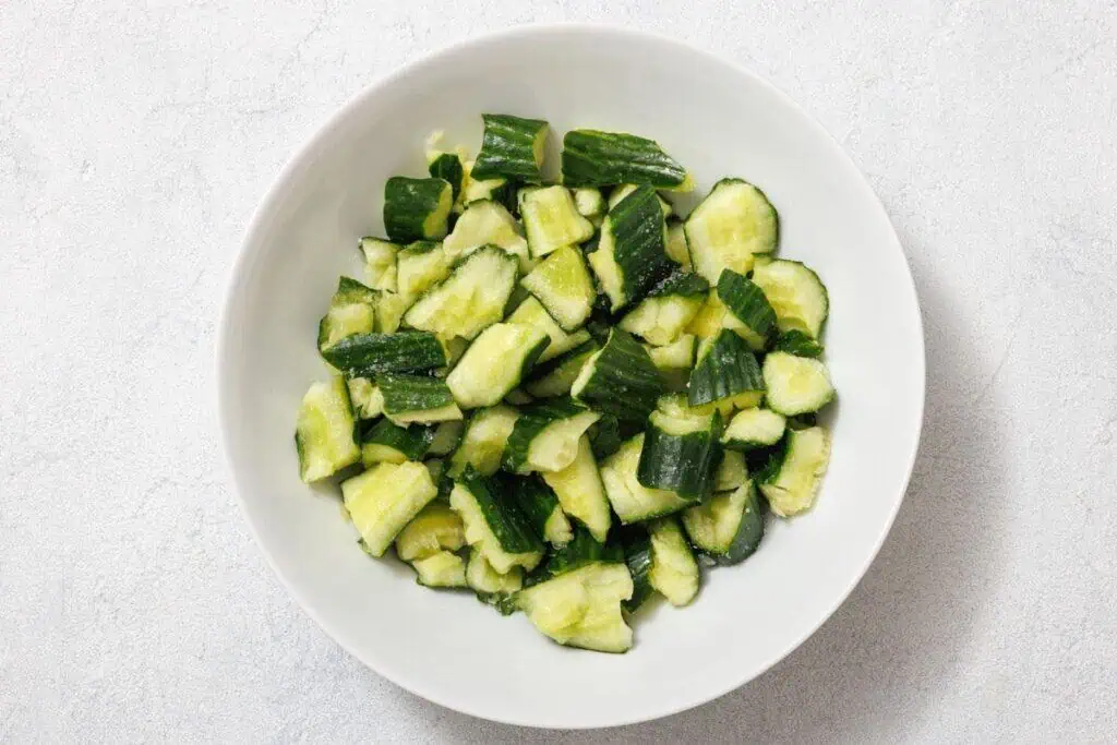 A white bowl filled with chopped cucumber pieces on a light gray surface.