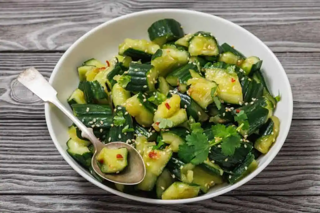 A bowl of cucumber salad with sesame seeds, cilantro, and red pepper flakes, served with a metal spoon on a wooden surface.
