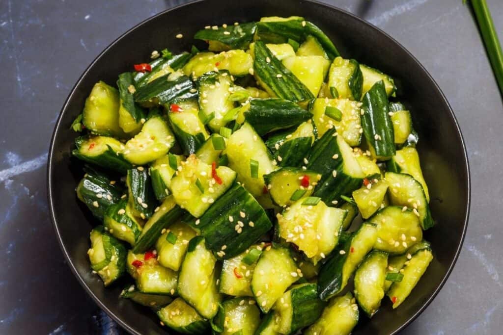 A bowl of sliced cucumbers tossed in a dressing with sesame seeds and chopped herbs, placed on a dark surface.