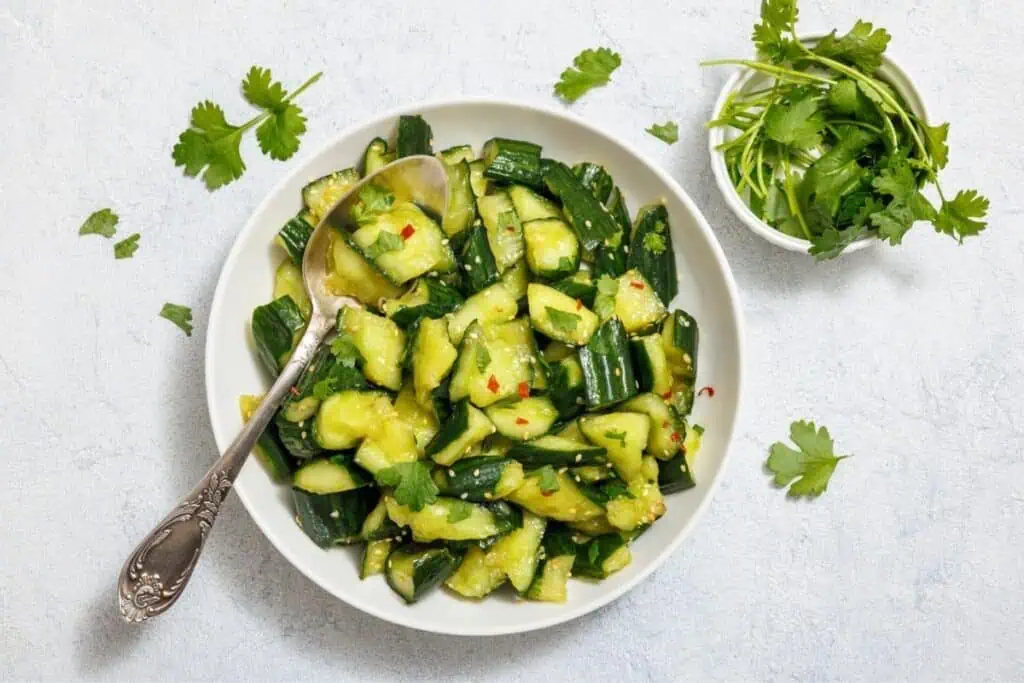 A white bowl of chopped cucumber salad with herbs and a spoon. A small bowl of fresh cilantro is beside it, with cilantro leaves scattered on a light background.