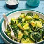 A bowl of cucumber salad with sesame seeds and herbs, accompanied by a fork. There's a small dish of salt and a ceramic jug in the background on a blue wooden table.