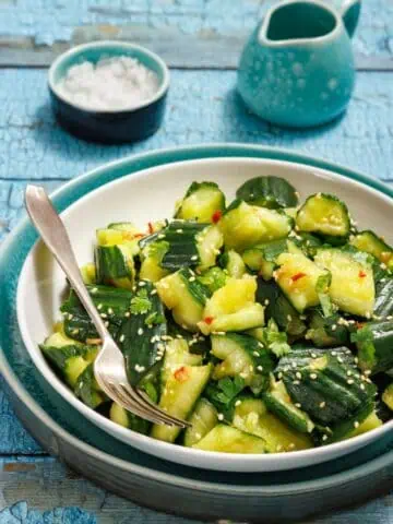 A bowl of cucumber salad with sesame seeds and herbs, accompanied by a fork. There's a small dish of salt and a ceramic jug in the background on a blue wooden table.