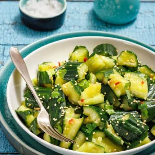 A bowl of cucumber salad with sesame seeds and herbs, accompanied by a fork. There's a small dish of salt and a ceramic jug in the background on a blue wooden table.