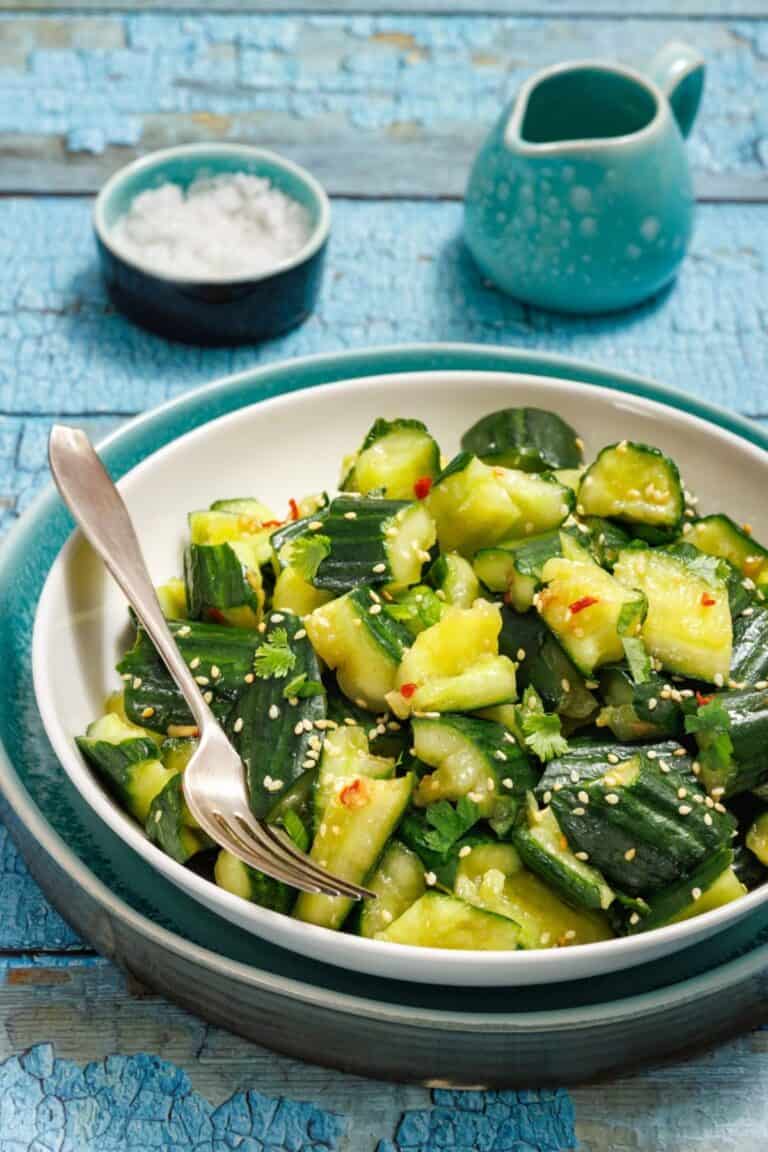 A bowl of cucumber salad with sesame seeds and herbs, accompanied by a fork. There's a small dish of salt and a ceramic jug in the background on a blue wooden table.