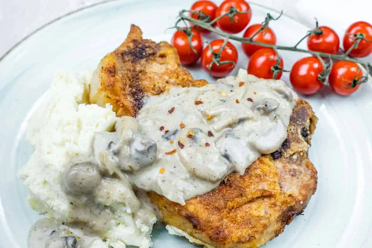 Plate with Cast Iron Smothered Pork Chops served alongside mashed potatoes and vine tomatoes.