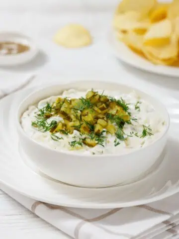 A bowl of creamy Dill Pickle Dip topped with chopped pickles and dill, placed on a white saucer. A plate of potato chips is in the background on a white tablecloth.