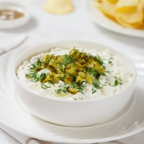 A bowl of creamy Dill Pickle Dip topped with chopped pickles and dill, placed on a white saucer. A plate of potato chips is in the background on a white tablecloth.