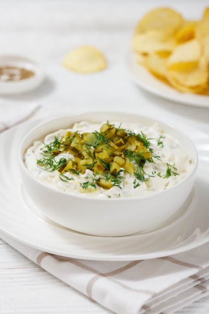 A bowl of creamy Dill Pickle Dip topped with chopped pickles and dill, placed on a white saucer. A plate of potato chips is in the background on a white tablecloth.