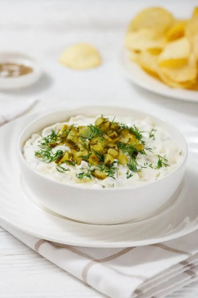 A bowl of creamy Dill Pickle Dip topped with chopped pickles and dill, placed on a white saucer. A plate of potato chips is in the background on a white tablecloth.
