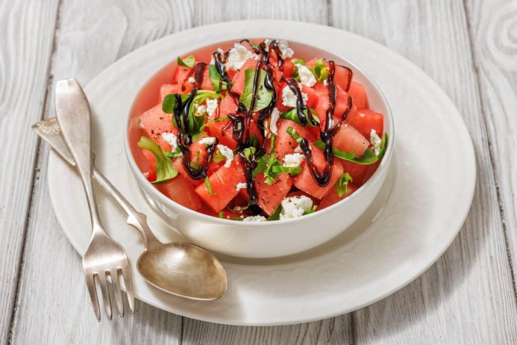 A bowl of watermelon salad with feta, basil, and a drizzle of balsamic glaze. Accompanied by a fork and spoon on a white plate, placed on a light wooden surface.