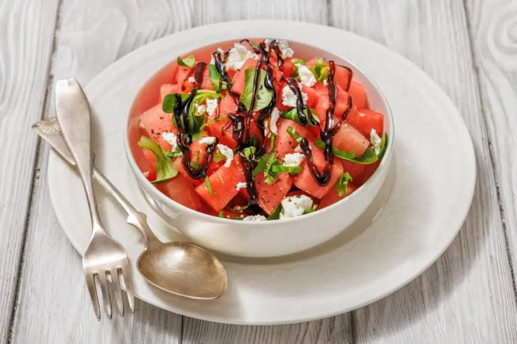 A bowl of watermelon salad with feta, basil, and a drizzle of balsamic glaze. Accompanied by a fork and spoon on a white plate, placed on a light wooden surface.