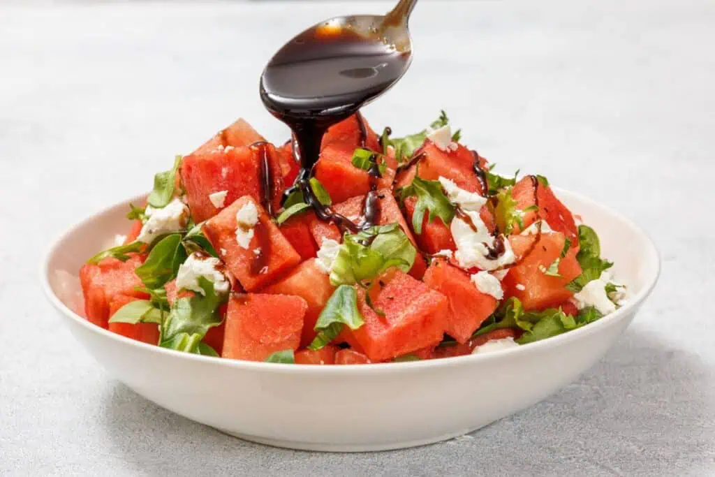 A bowl of watermelon salad with feta and greens is drizzled with balsamic glaze from a spoon.