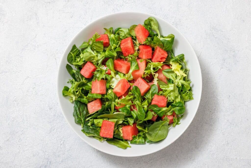 A white bowl contains a salad with mixed greens and cubes of watermelon on a light textured background.