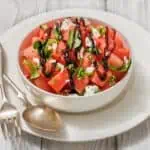 A bowl of watermelon salad with feta, basil, and a drizzle of balsamic glaze. Accompanied by a fork and spoon on a white plate, placed on a light wooden surface.