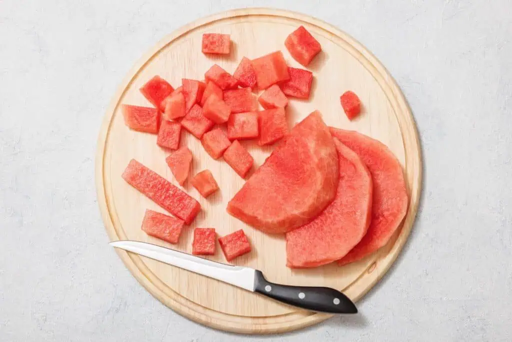 Cubed and sliced watermelon on a round wooden cutting board with a knife.
