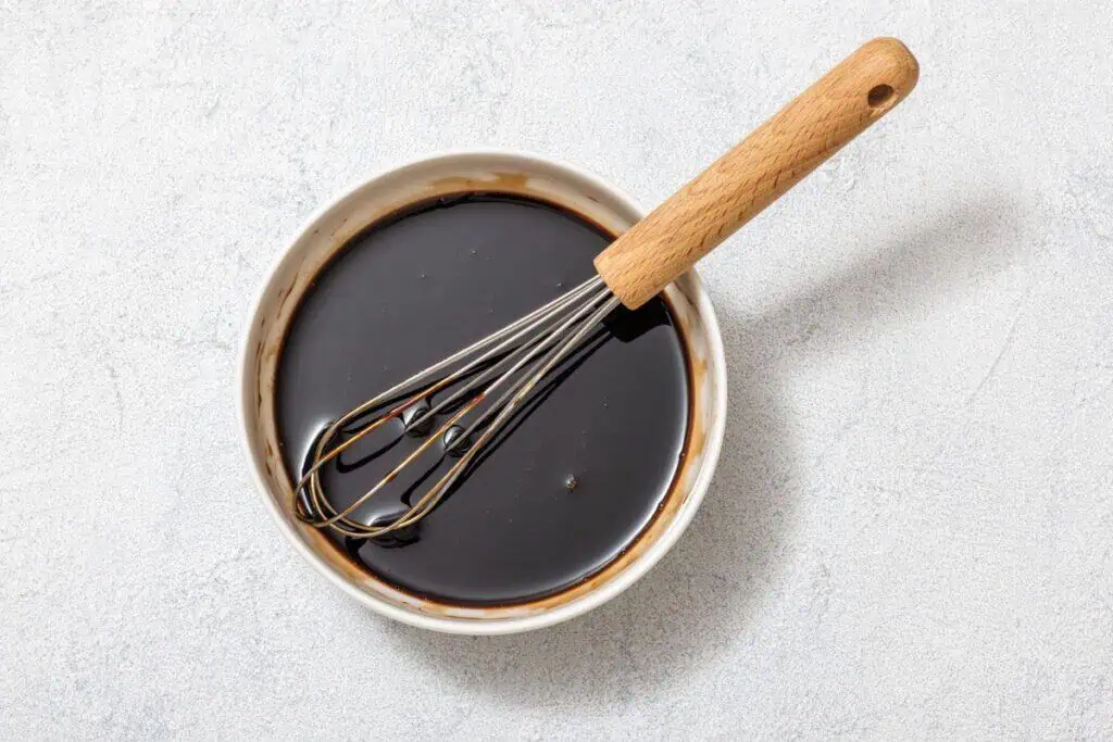 A whisk in a bowl of dark liquid on a light surface.