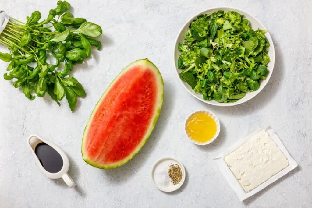 Ingredients for a watermelon salad: half a watermelon, a bowl of green leaves, feta cheese, honey, black pepper, balsamic vinegar, and fresh greens on a light surface.