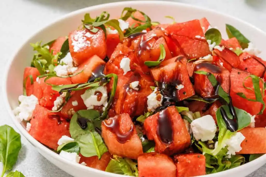 A bowl of watermelon salad with feta cheese, fresh greens, and a drizzle of balsamic glaze.