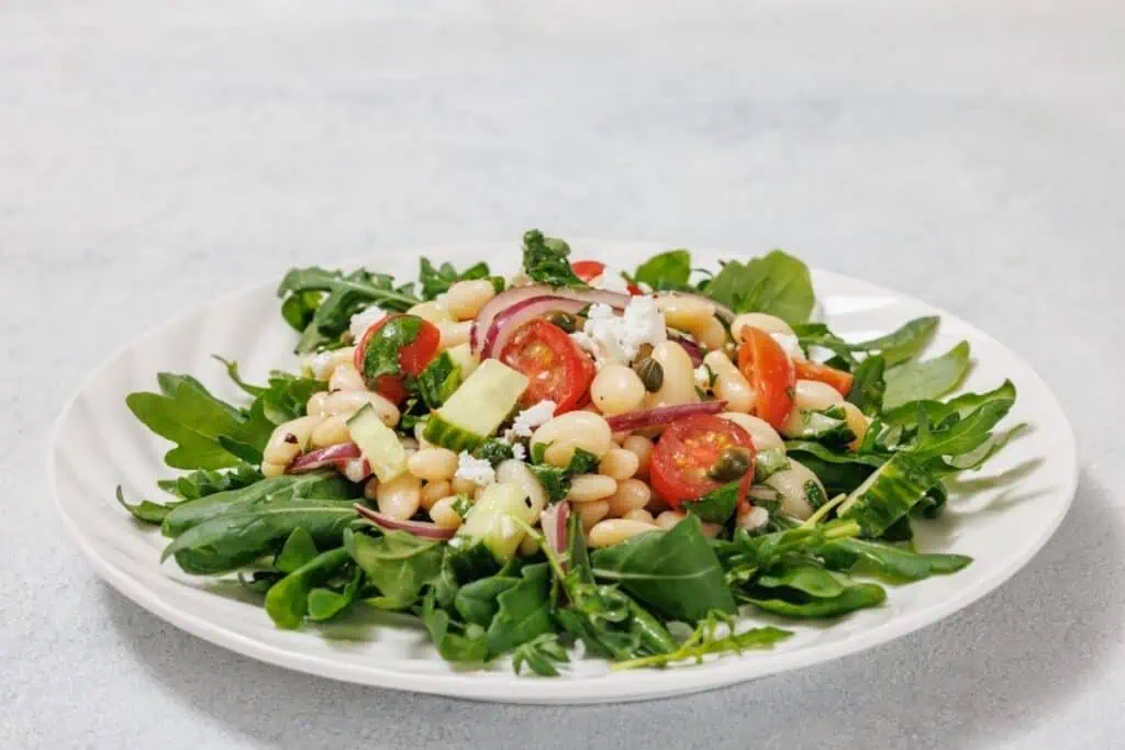 A White Bean Salad on a plate with arugula, cherry tomatoes, white beans, diced cucumber, red onion, and crumbled feta cheese.