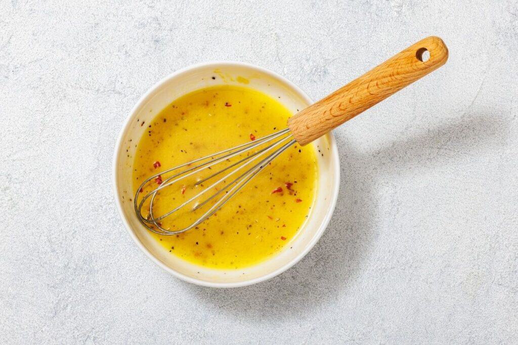 A bowl of yellow salad dressing with red flakes and a wooden-handled whisk on a light textured surface.