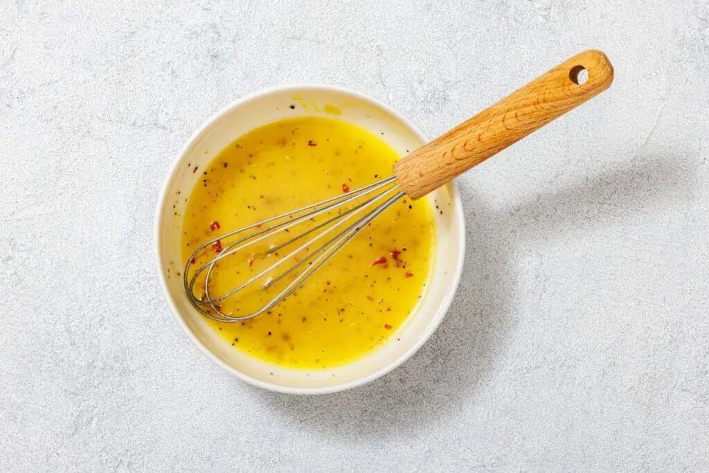 A bowl of yellow salad dressing with red flakes and a wooden-handled whisk on a light textured surface.
