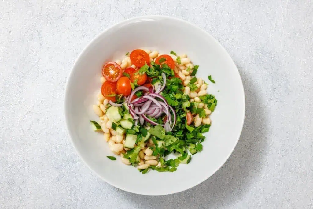 White bowl with a salad of white beans, cherry tomatoes, sliced red onions, cucumber, and chopped greens on a light gray background.