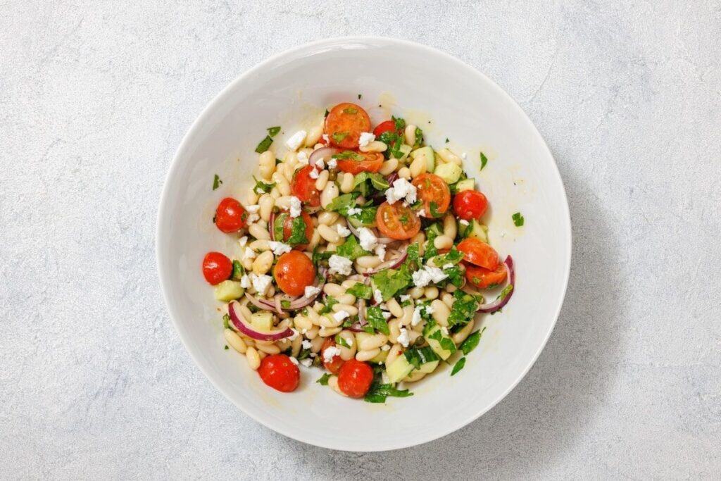 A bowl of salad with white beans, cherry tomatoes, cucumber, red onions, feta cheese, and herbs on a light gray background.