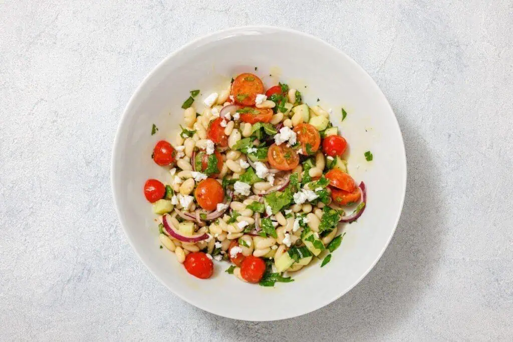A bowl of salad with white beans, cherry tomatoes, cucumber, red onions, feta cheese, and herbs on a light gray background.