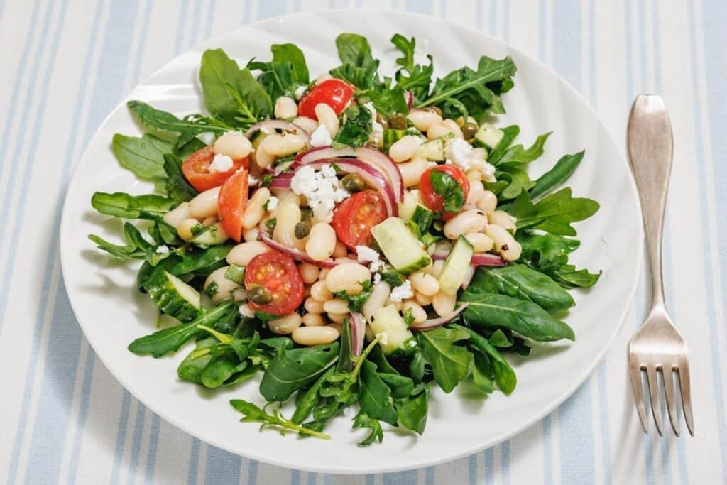 A white plate with a White Bean Salad featuring arugula, white beans, cherry tomatoes, red onions, cucumber, and crumbled cheese on a striped tablecloth, with a fork beside it.