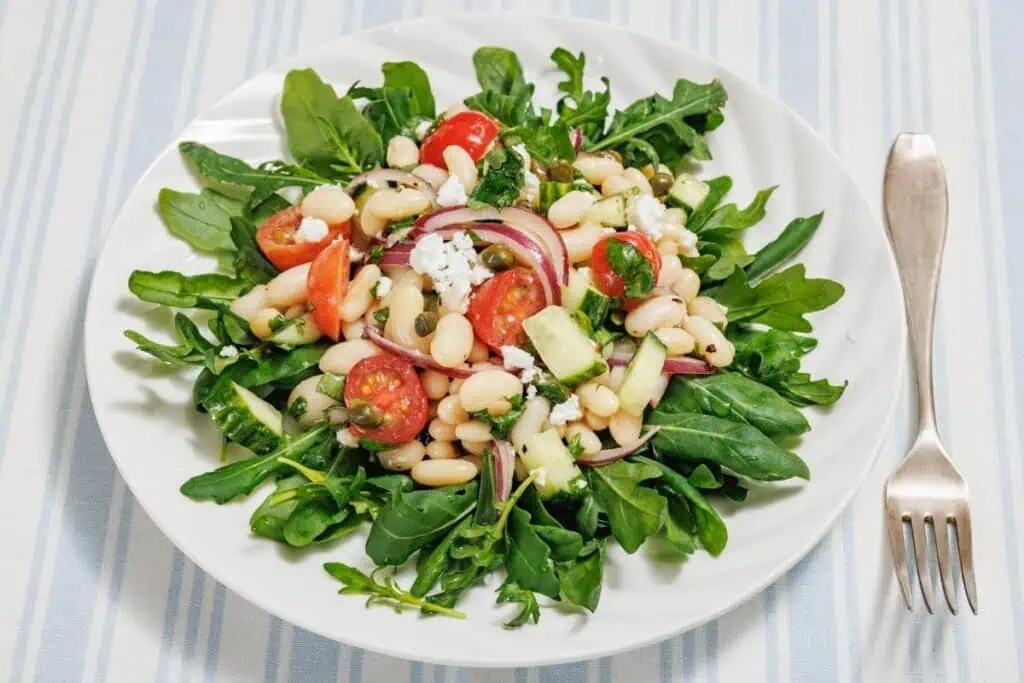 A white plate with a White Bean Salad featuring arugula, white beans, cherry tomatoes, red onions, cucumber, and crumbled cheese on a striped tablecloth, with a fork beside it.