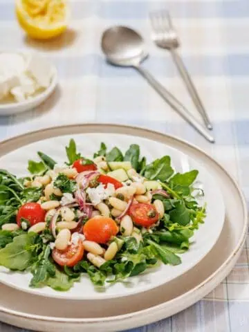A White Bean Salad featuring arugula, cherry tomatoes, white beans, and red onions on a plate. A cut lemon, feta, and utensils are in the background on a checkered tablecloth.