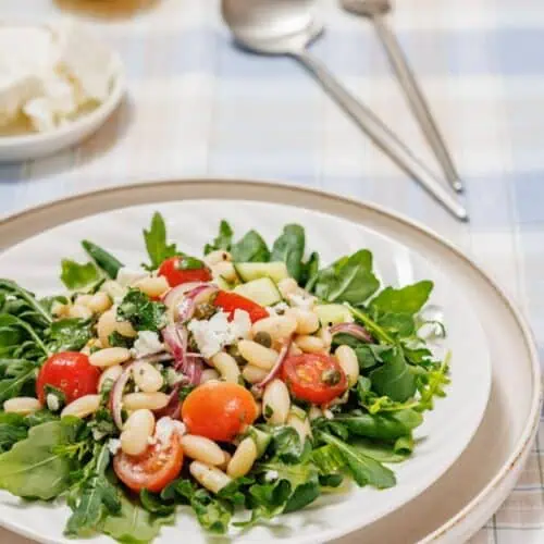 A White Bean Salad featuring arugula, cherry tomatoes, white beans, and red onions on a plate. A cut lemon, feta, and utensils are in the background on a checkered tablecloth.