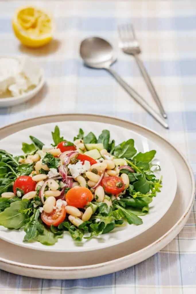A White Bean Salad featuring arugula, cherry tomatoes, white beans, and red onions on a plate. A cut lemon, feta, and utensils are in the background on a checkered tablecloth.