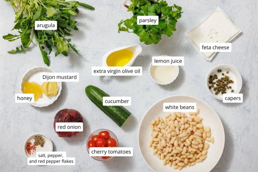Ingredients for White Bean Salad arranged on a table: arugula, parsley, feta cheese, capers, lemon juice, extra virgin olive oil, Dijon mustard, honey, cucumber, white beans, red onion, cherry tomatoes, spices.