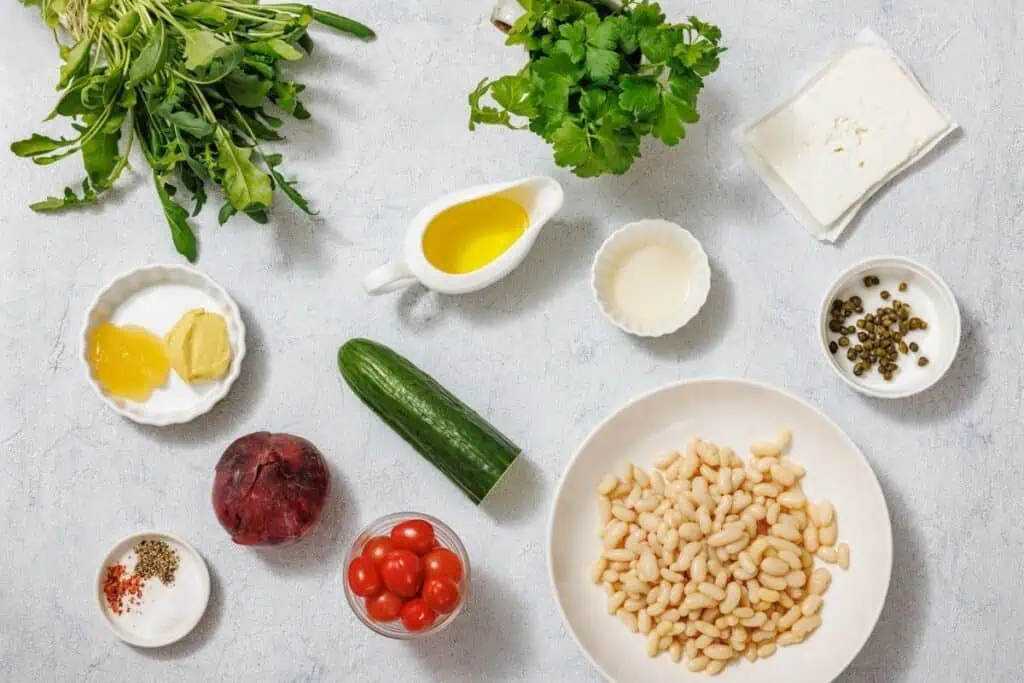 Ingredients for White Bean Salad arranged on a light surface: herbs, feta, olive oil, mustard, cream, capers, white beans, cucumber, cherry tomatoes, a beetroot, and spices.