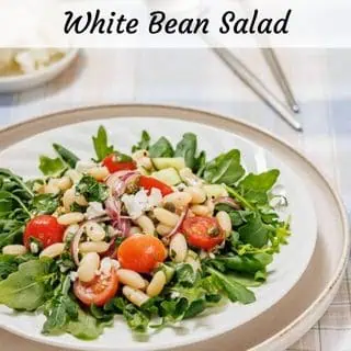 A plate of white bean salad with cherry tomatoes, arugula, red onions, and crumbled cheese on a checkered tablecloth.