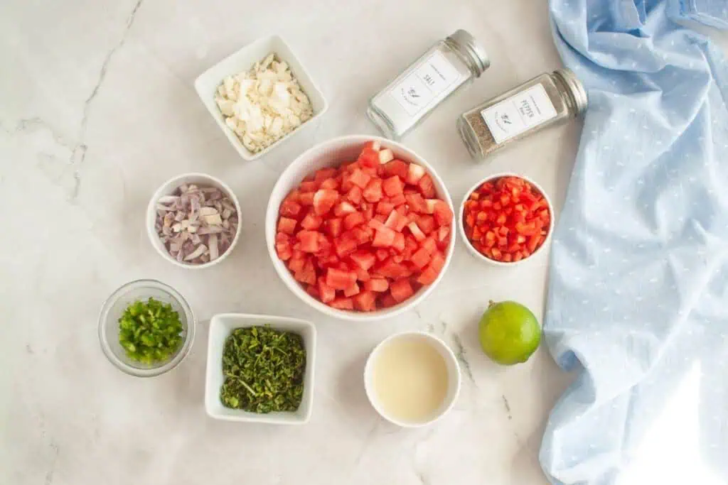 Overhead shot of ingredients for watermelon salad laid out in small bowls on a white marble surface, including diced watermelon, red bell pepper, shallots, feta cheese, jalapeno, cilantro, lime juice, lime, salt, and pepper. A blue cloth is draped on the side.