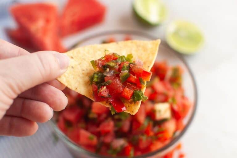 A hand holds a tortilla chip topped with red salsa, with a bowl of salsa, watermelon slices, and halved limes visible in the background.