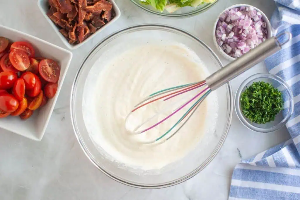 Overhead shot of a glass bowl containing mixed salad dressing with a whisk inside, surrounded by bowls of bacon, lettuce, red onion, parsley, and tomatoes on a marble surface.