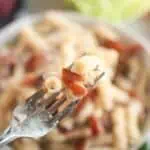 A close-up shot of a fork lifting pasta and bacon, with a blurry bowl of pasta salad in the background.
