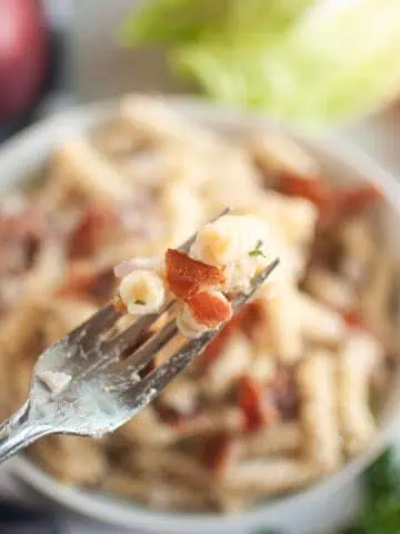 A close-up shot of a fork lifting pasta and bacon, with a blurry bowl of pasta salad in the background.