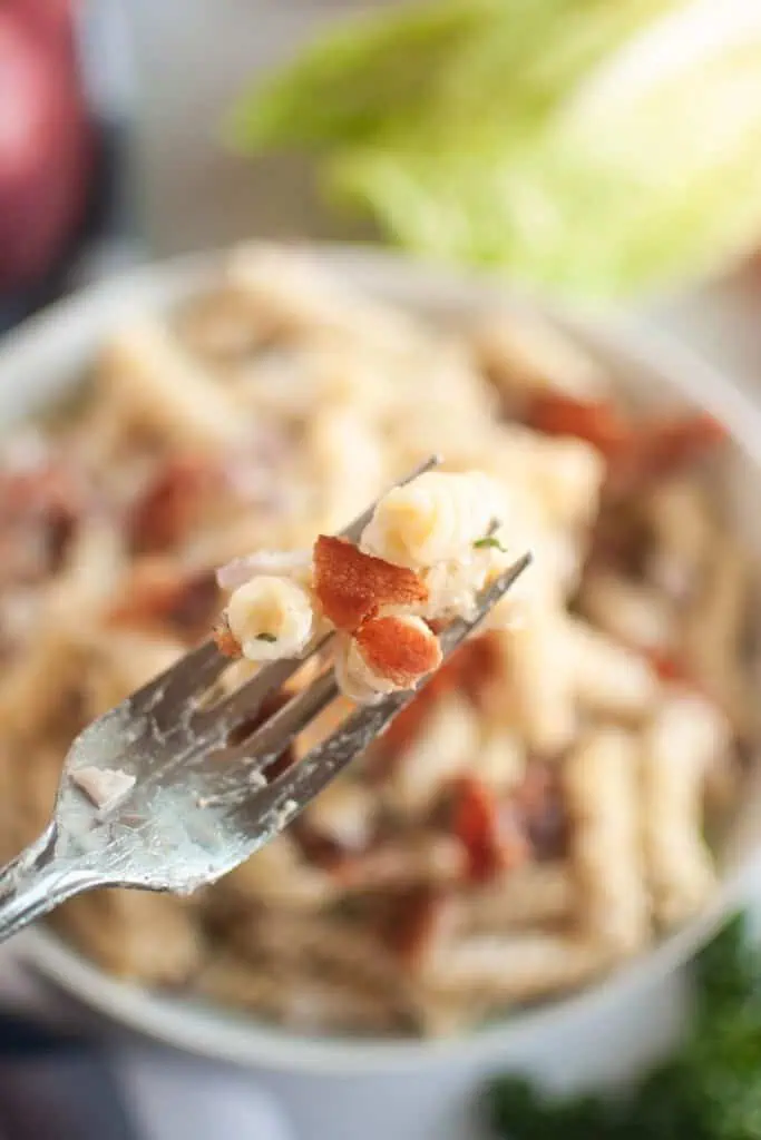 A close-up shot of a fork lifting pasta and bacon, with a blurry bowl of pasta salad in the background.