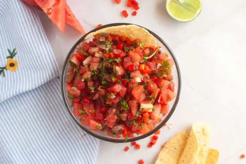 Overhead shot of a glass bowl filled with watermelon salsa, featuring diced watermelon, red bell pepper, and herbs. Tortilla chips and a lime half are visible around the bowl.