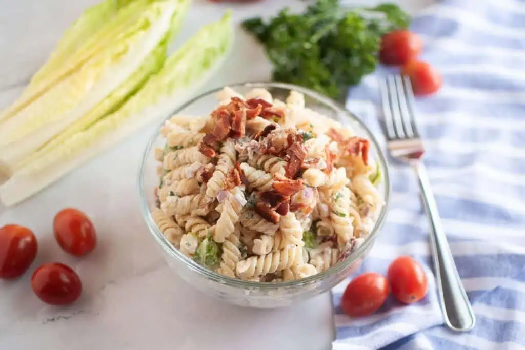 Overhead shot of a glass bowl filled with pasta salad, featuring spiral pasta, creamy dressing, pieces of bacon, and other visible ingredients. Lettuce, tomatoes, and parsley are arranged around the bowl.
