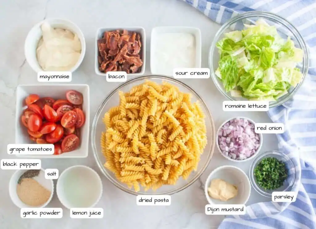 Overhead shot of various ingredients for a pasta salad, including cooked spiral pasta in a glass bowl, bowls of bacon, lettuce, red onion, parsley, tomatoes, and dressing components, arranged on a marble surface.