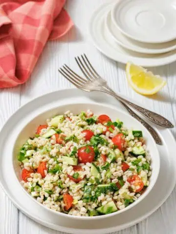 A bowl of barley salad with cherry tomatoes, cucumber, herbs, and greens, placed on a white plate with forks, lemon wedges, and a red napkin on a white wooden table.
