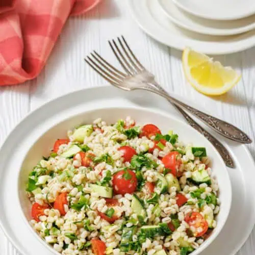A bowl of barley salad with cherry tomatoes, cucumber, herbs, and greens, placed on a white plate with forks, lemon wedges, and a red napkin on a white wooden table.