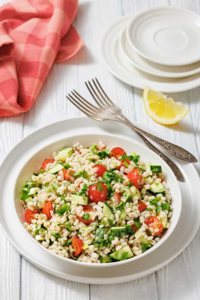 A bowl of barley salad with cherry tomatoes, cucumber, herbs, and greens, placed on a white plate with forks, lemon wedges, and a red napkin on a white wooden table.