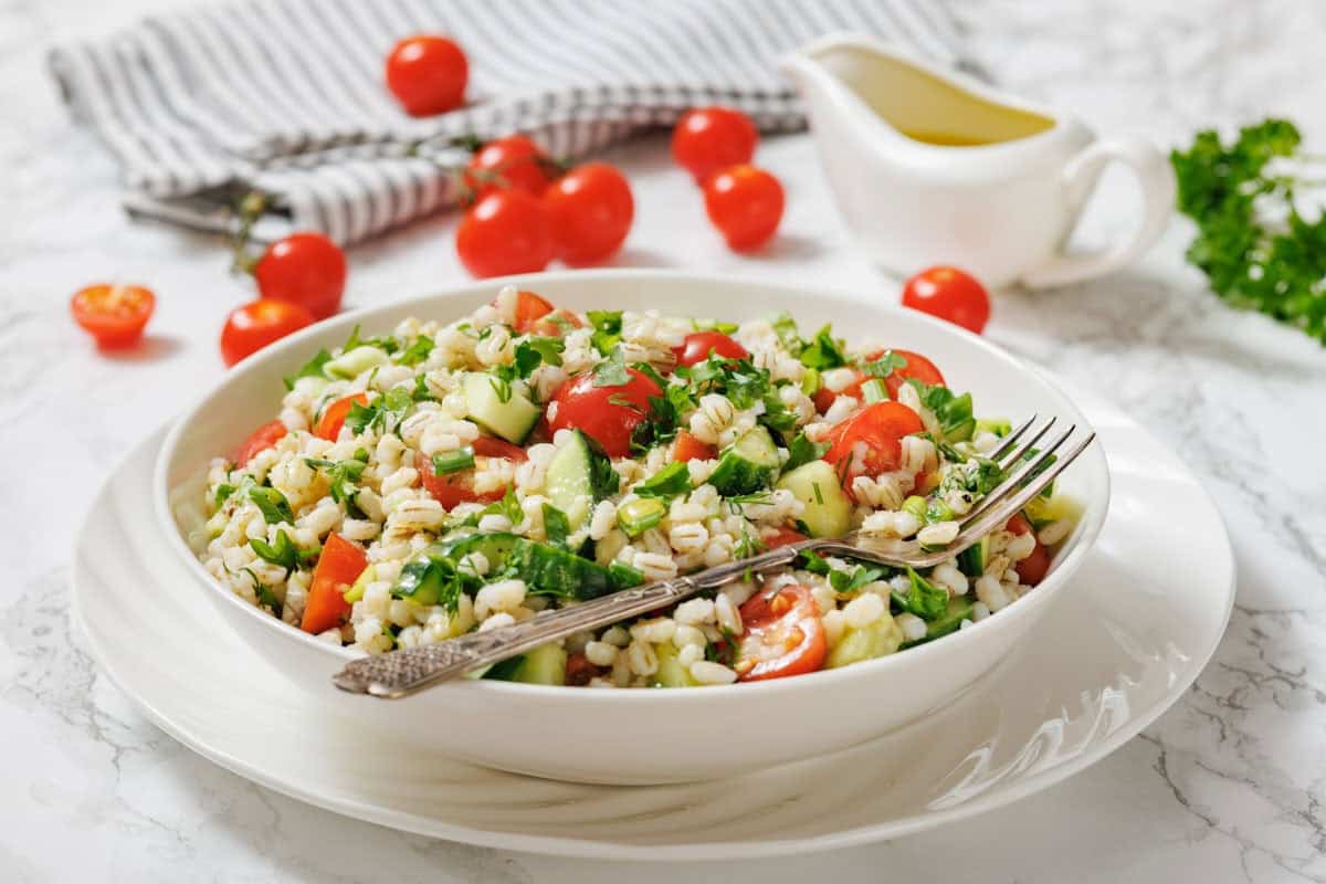 A bowl of barley salad with chopped tomatoes, cucumbers, parsley, and green onions, served with a fork. Olive oil and fresh cherry tomatoes are in the background.