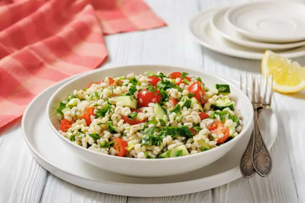 A bowl of Mediterranean-Inspired Barley Salad with cherry tomatoes, cucumber, and herbs sits on a plate beside forks, with a lemon wedge and folded napkin in the background.